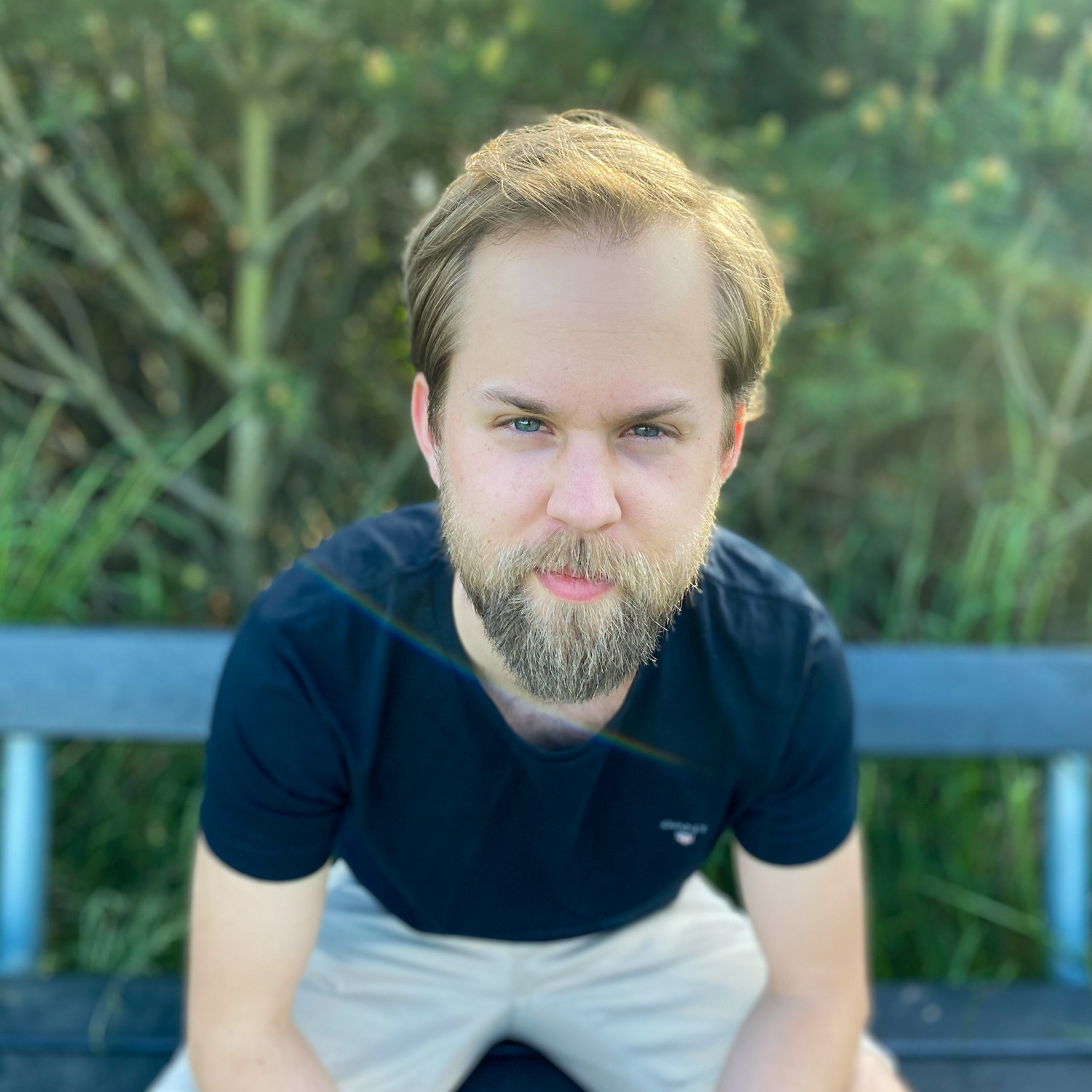 Markus sitter framåtlutad på en bänk på en bänk och ser in i kameran. Bakom bänken finns en grönskande buske och man kan se att solen lyser genom löven.